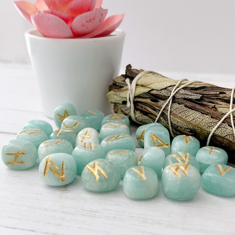 May include: A set of 24 teal green tumbled stones with gold runes inscribed on each. The stones are arranged in a loose pile on a white surface.
