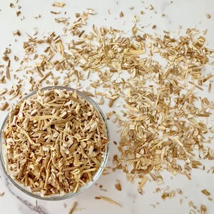 May include: A clear glass bowl filled with light brown, chopped pieces of a root, with more of the same scattered around it. The pieces are of varying lengths and widths, and the background is a white surface.