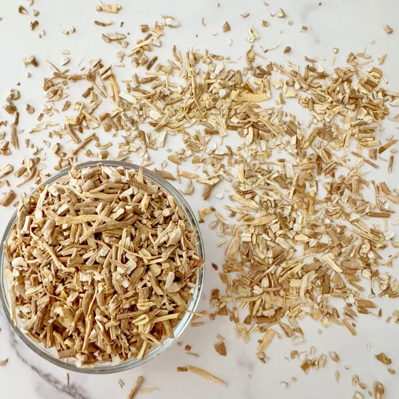 May include: A clear glass bowl filled with light brown, chopped pieces of a root, with more of the same scattered around it. The pieces are of varying lengths and widths, and the background is a white surface.