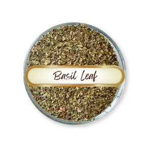 May include: A circular glass bowl filled with dried, finely chopped green and brown basil leaves. A horizontal, cream-colored rectangular label with a gold border is placed across the center, featuring the text Basil Leaf in dark brown script.