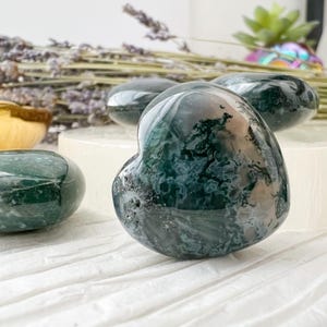 May include: Several polished green moss agate heart-shaped stones. The stones have a translucent appearance with dark green and white patterns. The stones are displayed on a white surface with dried lavender and a small gold-colored bowl in the background.