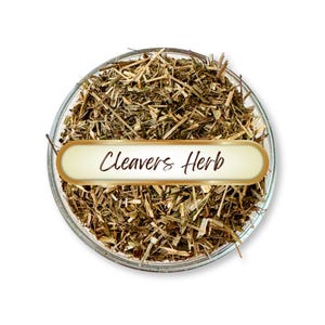 May include: A clear glass bowl filled with dried, chopped herbs. A gold-edged label with the words "Cleavers Herb" is centered in the bowl. The herbs are a mix of green and brown.