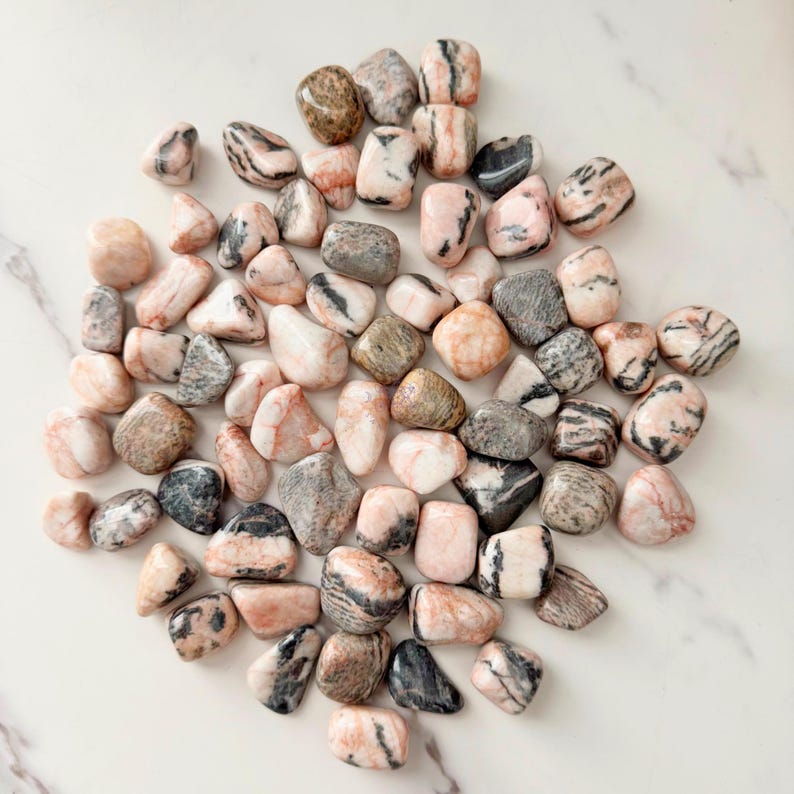 May include: A collection of tumbled stones in shades of pink, gray, and brown. The stones vary in size and shape, with some displaying striped patterns. The polished surfaces reflect light, creating a natural and earthy aesthetic.