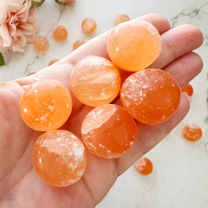 May include: A collection of seven orange carnelian spheres, each with a unique pattern of white veins and swirls. The spheres are smooth and polished, and they are arranged in a hand.