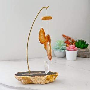 May include: A decorative wind chime featuring orange agate slices suspended from a gold-colored metal stand. The base is a natural stone with a polished top. A clear crystal hangs below the agate slices. Decorative succulents are in the background.