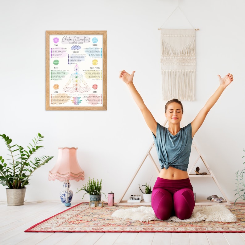 Op de afbeelding: Een vrouw zit in een yogahouding met haar armen omhoog. Achter haar, aan de muur, hangt een ingelijst poster met de titel "Chakra Affirmations" en een kleurrijk chakra-diagram. De poster somt de zeven chakra's op en hun bijbehorende kleuren, elementen en affirmaties.