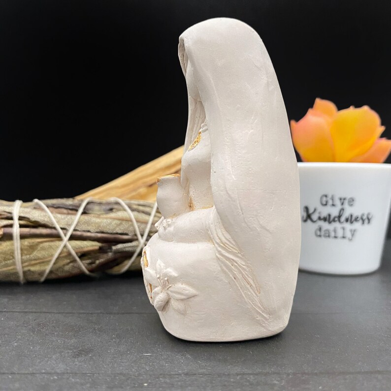 Puede incluir: Estatua de color crema de una figura sentada con la cabeza cubierta con un velo, sosteniendo un peque&ntilde;o recipiente. La estatua tiene detalles dorados y florales en la base. Un manojo de salvia y una maceta con el texto "Give Kindness daily" est&aacute;n en el fondo.