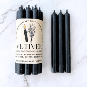 May include: A bundle of black spell candles with a Vetiver label, alongside a separate group of black candles. The label features a drawing of a candle and a plant, with text that reads "VETIVER" and "THE STONES SANCTUARY SHOP ESTABLISHED".