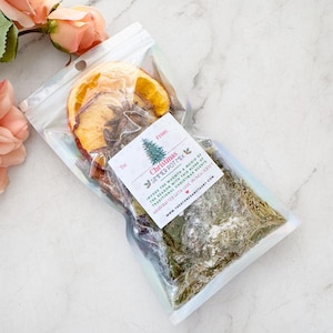 May include: A clear plastic bag filled with dried orange slices, cinnamon sticks, and herbs. A white label reads "Christmas Simmer Pot Mix" with text about traditional Christmas scents. The bag is sealed and ready for use.