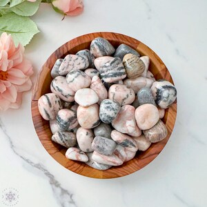 May include: A wooden bowl filled with polished stones. The stones are a mix of pink, gray, and white, with unique striped patterns. The bowl sits on a white marble surface, with pink flowers in the background.