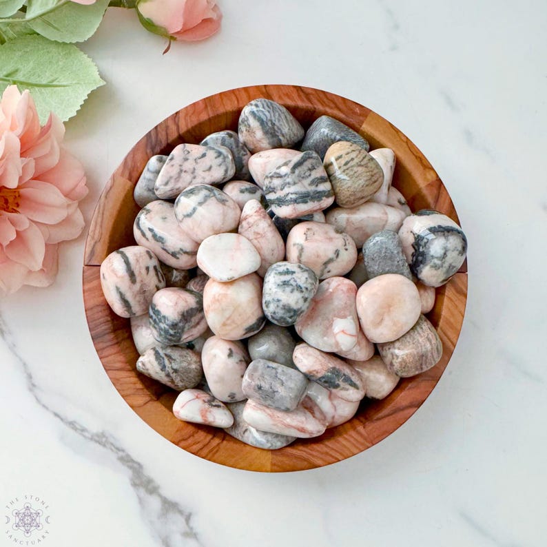May include: A wooden bowl filled with polished stones. The stones are a mix of pink, gray, and white, with unique striped patterns. The bowl sits on a white marble surface, with pink flowers in the background.