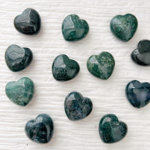 May include: A collection of polished heart-shaped stones in varying shades of green. The stones have a glossy finish and appear to be made of a natural material. The hearts are arranged on a white, textured surface.