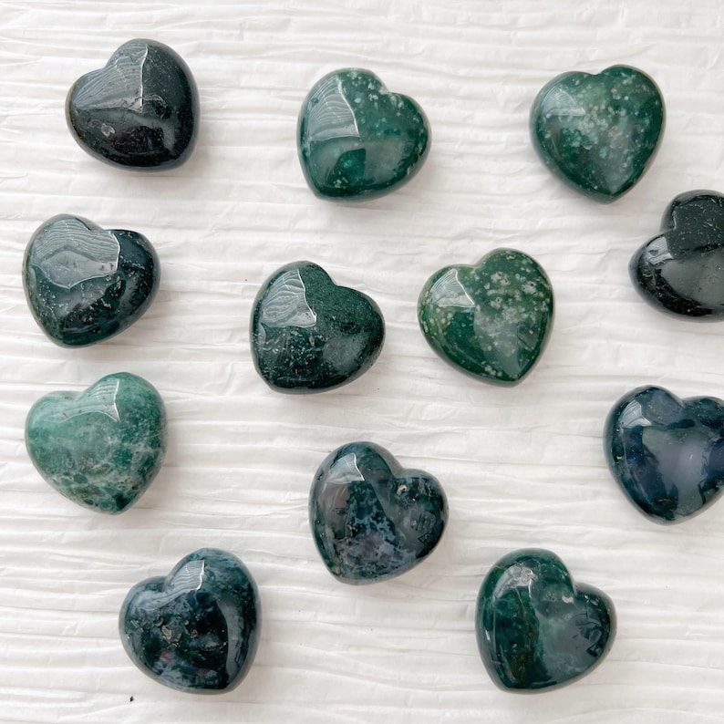May include: A collection of polished heart-shaped stones in varying shades of green. The stones have a glossy finish and appear to be made of a natural material. The hearts are arranged on a white, textured surface.