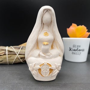Puede incluir: Estatua de color crema de una figura sentada con una larga cobertura para la cabeza. La figura sostiene un peque&ntilde;o recipiente y lleva un colgante dorado. La base presenta un dise&ntilde;o floral dorado. Una peque&ntilde;a maceta con el texto "Give Kindness daily" est&aacute; en el fondo.