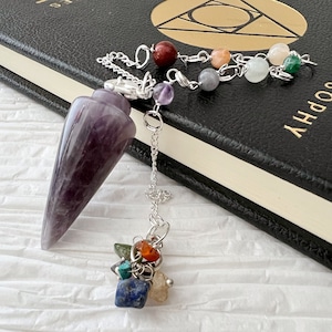 May include: Amethyst pendulum with a silver chain and a cluster of colorful crystals at the bottom. The pendulum is resting on a black book with the word "SOPHIA" printed on the cover.