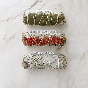 May include: Three smudge sticks, tied with white string, are arranged vertically on a white marble surface. The top stick is green and labeled "Basil + Sage". The middle stick is white with red flowers and labeled "Red Sinuata + Sage". The bottom stick is white and labeled "White Sage Stick".