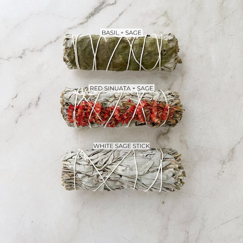 May include: Three smudge sticks, tied with white string, are arranged vertically on a white marble surface. The top stick is green and labeled "Basil + Sage". The middle stick is white with red flowers and labeled "Red Sinuata + Sage". The bottom stick is white and labeled "White Sage Stick".