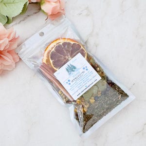 May include: A clear plastic bag filled with a simmer pot mix, including dried orange slices, cinnamon sticks, and various herbs and spices. The bag has a label that reads "Winter Simmer Pot Mix" and includes the text "Welcome the stillness of winter with a simmering blend that captures the essence of cozy days & fireside nights. Handcrafted with love in New Jersey. www.thestonesanctuary.com".