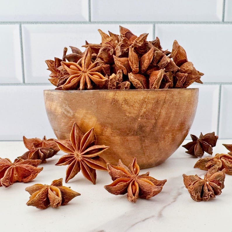 K&ouml;nnte beinhalten: Eine Holzschale gef&uuml;llt mit Sternanis, mit mehreren St&uuml;cken darum verstreut. Der Sternanis hat eine tiefrotebraune Farbe, und die Schale hat einen warmen, nat&uuml;rlichen Holzton. Der Hintergrund ist eine wei&szlig;e Fliesenoberfl&auml;che.
