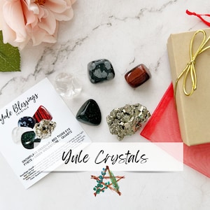 May include: A set of Yule crystals in a gift box. The crystals include snowflake obsidian, red tiger eye, pyrite, green goldstone, and clear quartz. The box is wrapped in red tissue paper and tied with a gold ribbon.