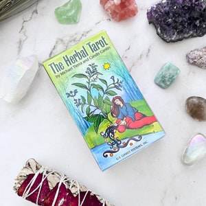 May include: A deck of "The Herbal Tarot" cards with a colorful illustration of a woman and plants. The box is surrounded by crystals and a sage bundle. The text on the box reads "The Herbal Tarot" by Michael Tierra and Candis Cantin.