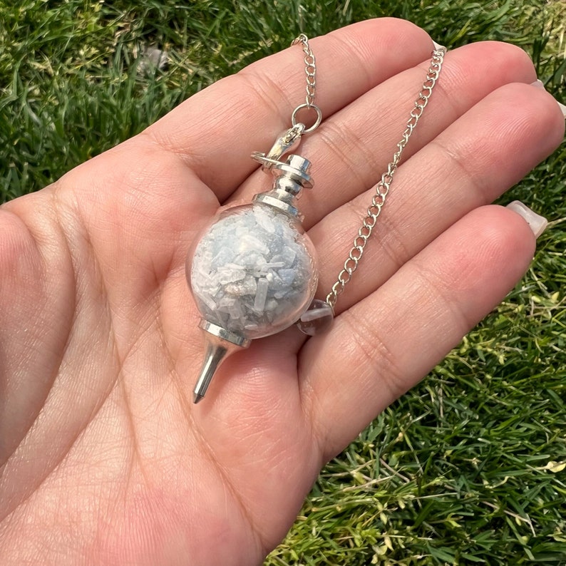 May include: A silver pendulum with a clear glass sphere filled with white selenite crystals. The pendulum is attached to a silver chain.
