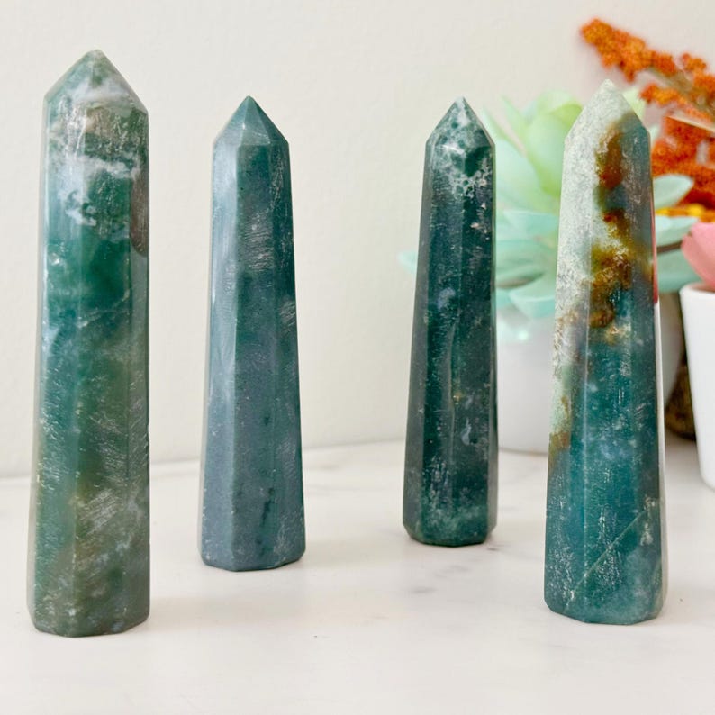 May include: Four green moss agate crystal towers with pointed tops. The crystals have a mix of green and white patterns. The towers are standing upright, showcasing their polished surfaces and natural variations. They are arranged on a white surface.