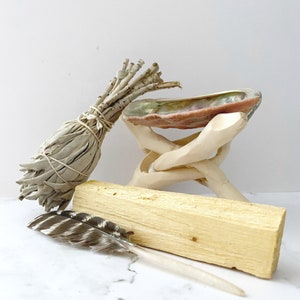 Puede incluir: Un manojo de salvia blanca, un palo santo, una pluma y una concha de mar sobre un soporte de madera. Los art&iacute;culos se utilizan para rituales de limpieza y purificaci&oacute;n.