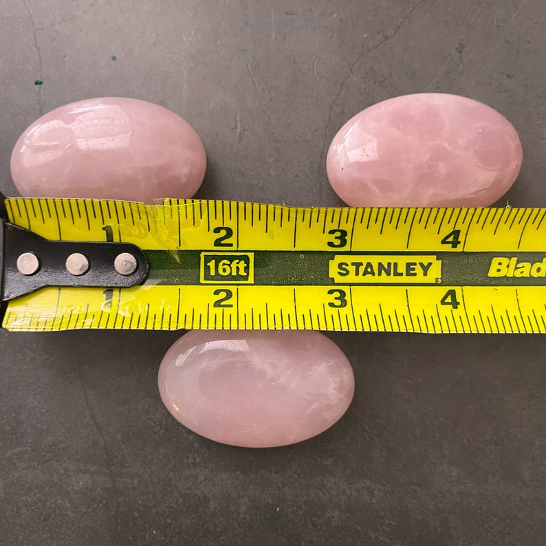 Pu&ograve; includere: Tre pietre di quarzo rosa di forma ovale rosa, ciascuna misura circa 5 cm di lunghezza, sono disposte su una superficie grigia. Un metro a nastro giallo &egrave; posizionato sotto le pietre, con la misura di 5 cm segnata.