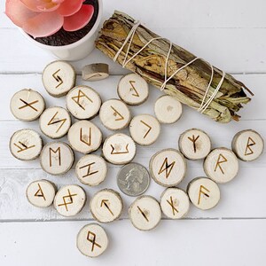 May include: A set of 25 wooden runes with Norse symbols carved into them. The runes are arranged on a white wooden surface with a bundle of dried sage in the background.
