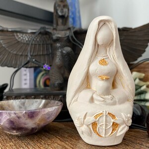 Puede incluir: Una estatua de color crema de una figura femenina sentada con un velo drapeado. La estatua tiene detalles dorados en el pecho, el vientre y la base, que presenta un dise&ntilde;o floral. La figura sostiene un peque&ntilde;o recipiente.