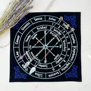 May include: A black velvet pendulum board with a white circle in the center and the days of the week and zodiac signs around the circle. Three silver chains with crystal pendulums are hanging over the board.