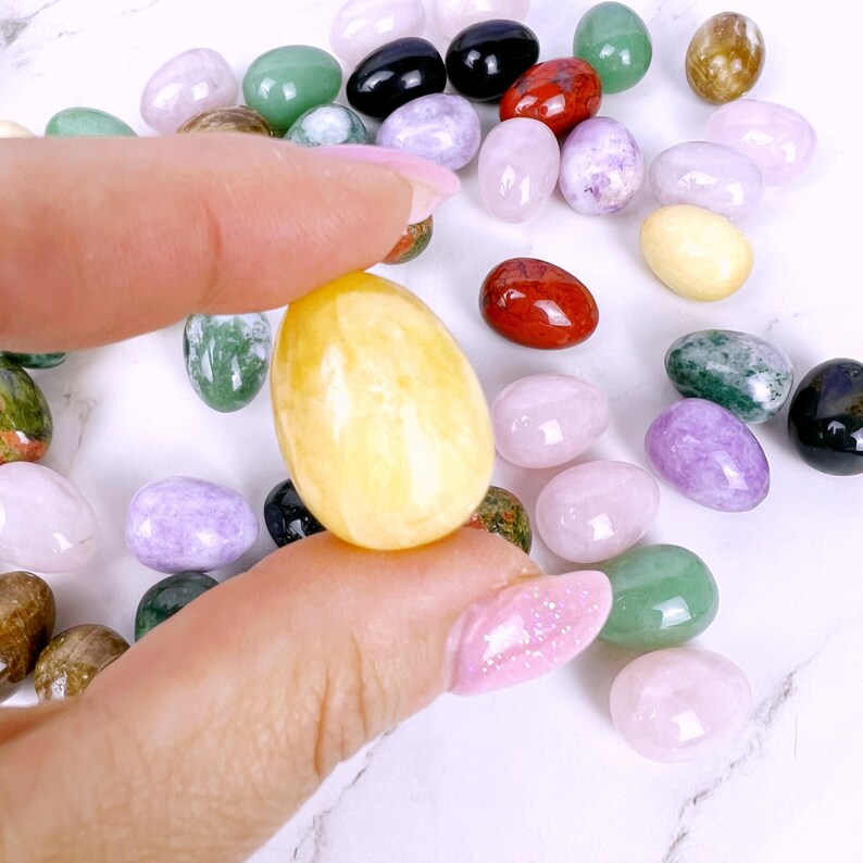 Puede incluir: Una colecci&oacute;n de huevos de piedras preciosas pulidas en varios colores, incluyendo verde, rojo, morado y amarillo. Los huevos est&aacute;n dispuestos sobre una superficie blanca.