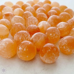 May include: A collection of orange carnelian spheres, each with a unique white marbling pattern. The spheres are smooth and polished, and they are arranged in a random pattern on a white surface.
