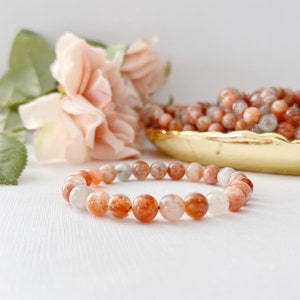 May include: A bracelet made of orange, white, and clear sunstone beads. The bracelet is on a white surface with a pink rose in the background.
