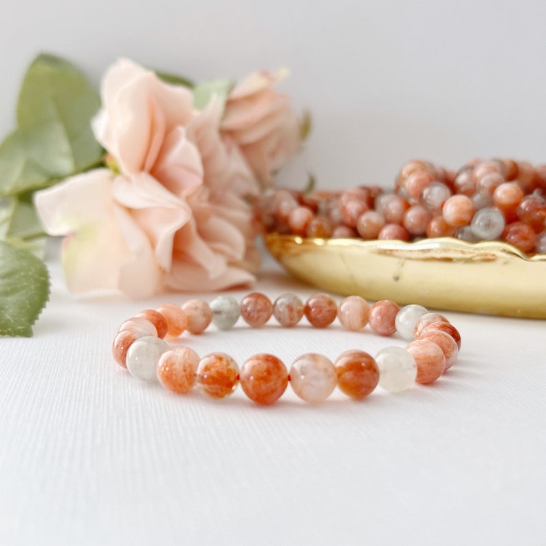 May include: A bracelet made of orange, white, and clear sunstone beads. The bracelet is on a white surface with a pink rose in the background.