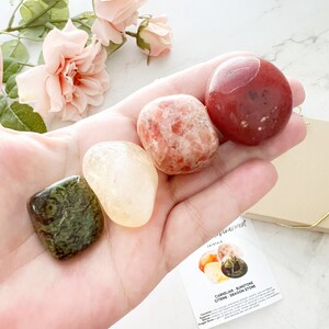 May include: A hand holding four tumbled stones: a green and brown dragon stone, a yellow citrine, a red carnelian, and an orange sunstone. The stones are arranged in a semi-circle on the palm of the hand. A card with the text "Carnelian, Sunstone, Citrine, Dragon Stone" is visible in the background.