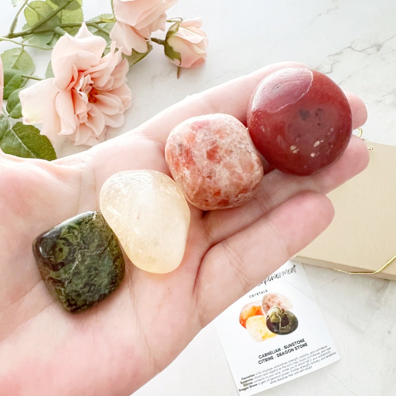 May include: A hand holding four tumbled stones: a green and brown dragon stone, a yellow citrine, a red carnelian, and an orange sunstone. The stones are arranged in a semi-circle on the palm of the hand. A card with the text "Carnelian, Sunstone, Citrine, Dragon Stone" is visible in the background.