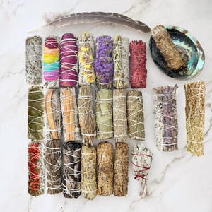 May include: Assortment of 20 smudge sticks, including white sage, lavender, cedar, and palo santo. The sticks are tied with twine and some have dried flowers or herbs incorporated into them. A feather and a seashell are also included in the image.
