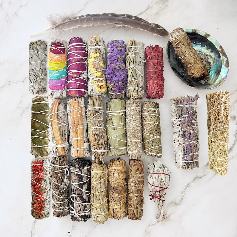 May include: Assortment of 20 smudge sticks, including white sage, lavender, cedar, and palo santo. The sticks are tied with twine and some have dried flowers or herbs incorporated into them. A feather and a seashell are also included in the image.