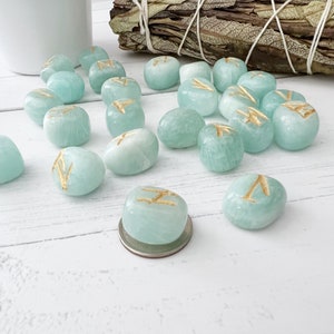 May include: A set of 15 teal green gemstone rune stones with gold runes etched on each stone. The stones are scattered on a white wooden surface.