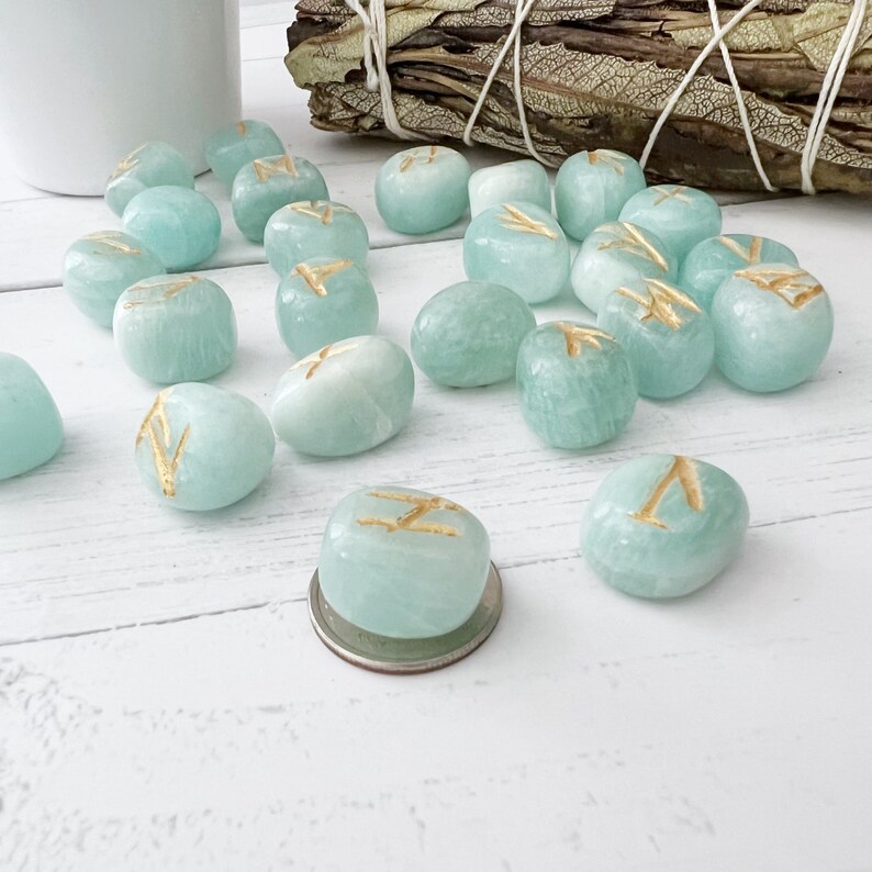 May include: A set of 15 teal green gemstone rune stones with gold runes etched on each stone. The stones are scattered on a white wooden surface.