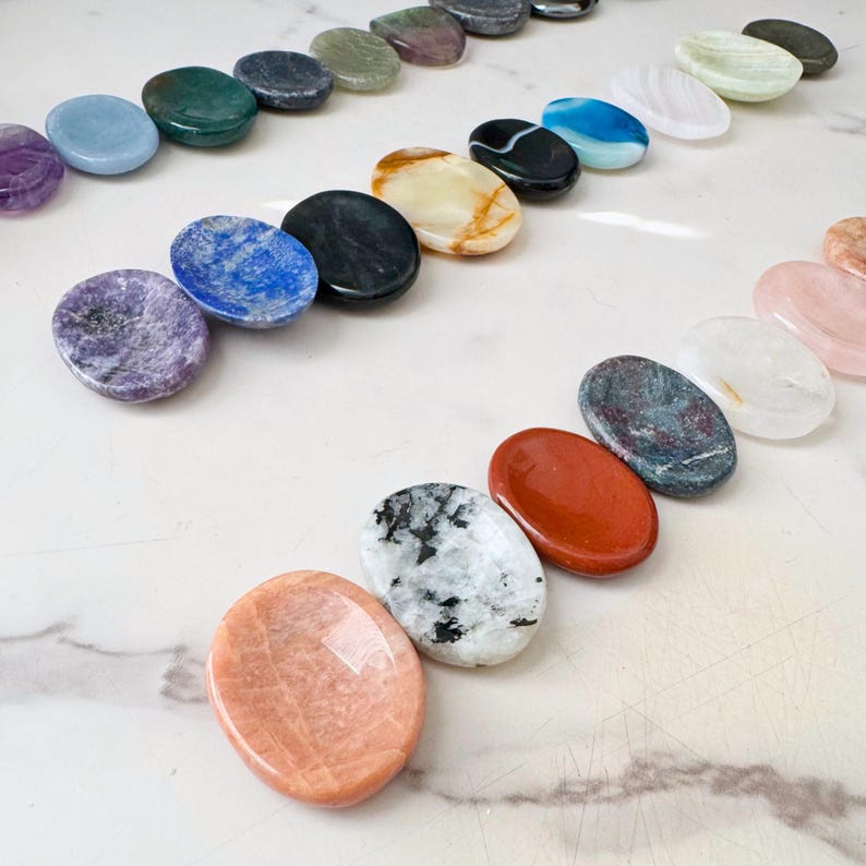 May include: Assortment of polished gemstones in various colors, including purple, blue, green, and orange. The stones are oval-shaped and arranged in rows, showcasing natural patterns and textures. Ideal for crystal healing or decorative purposes.
