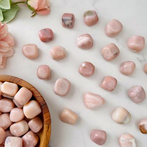 May include: A collection of polished pink gemstones scattered on a white surface, with some stones in a wooden bowl. The stones vary in shade from light to medium pink, with some showing natural patterns. A few pink flowers are visible in the background.