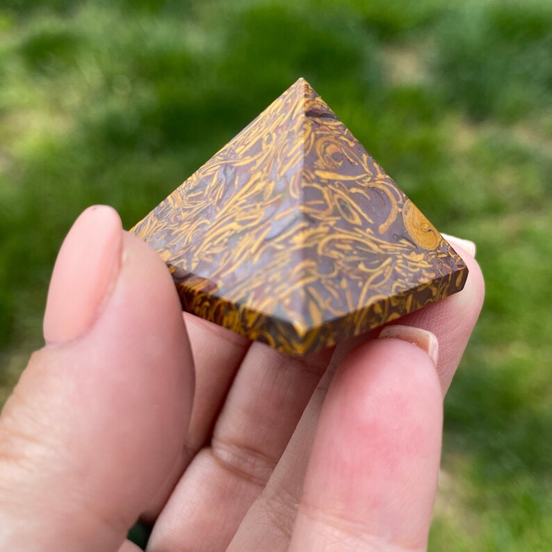May include: A pyramid-shaped decorative stone with a brown base and yellow, swirled patterns. The pyramid is held in a hand against a blurred green background.