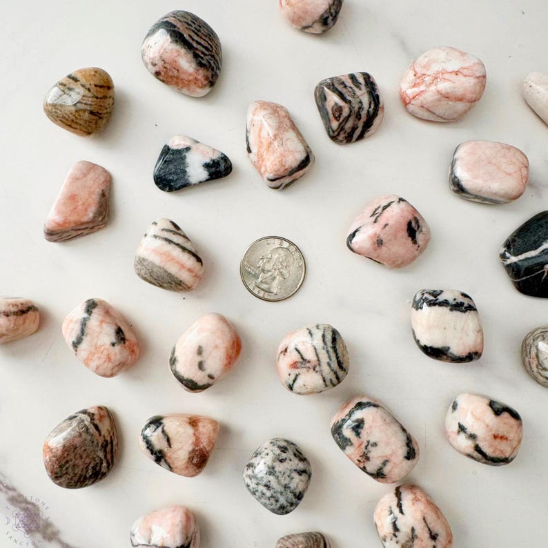 May include: A collection of polished stones in shades of pink, brown, black, and white, scattered on a white surface. A US quarter coin is included for size reference. The stones vary in shape and have unique patterns and textures.