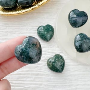 May include: Several polished heart-shaped stones in shades of green and white are displayed. The stones have a glossy finish and varying patterns. Some are on a gold-colored tray and a white plate, while others are held by a hand.