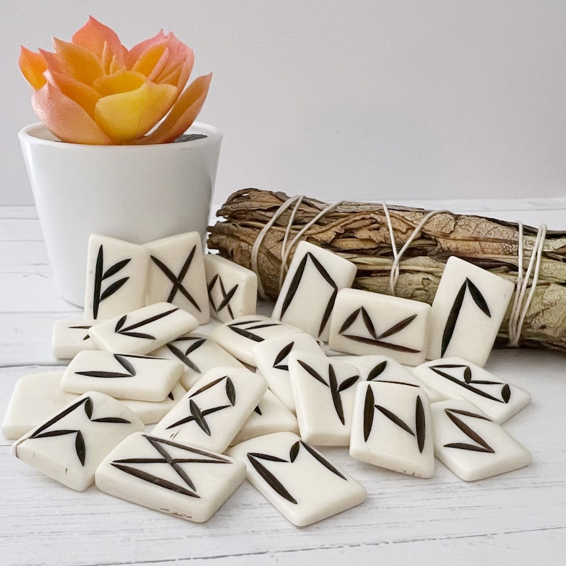 May include: A collection of off-white rune stones with black etched symbols. The stones are arranged on a white surface, next to a small white pot with an orange succulent and a bundle of dried sage.
