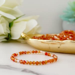 May include: A delicate bracelet made of small, round, orange and clear beads. The bracelet is displayed on a white surface, with a gold-colored dish filled with similar beads in the background. A white flower is also visible.