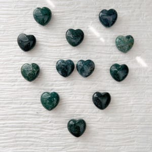 May include: A collection of heart-shaped stones in varying shades of green and dark teal. The stones have a polished, smooth surface and are arranged in a scattered pattern. The stones appear to be made of a natural material.